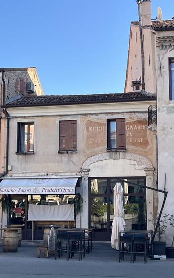 A rustic stone building with overhangs has taller structures on each side in Bassano del Grappa, Italy.