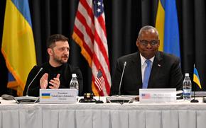 Ukrainian President Vlodymyr Zelenskyy claps as U.S. Defense Secretary Lloyd Austin smiles at a table with the Ukraine and U.S. flags in the background.  