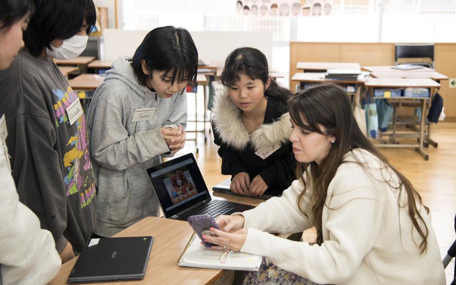 An American teacher sits at a desk and shows a phone to a group of Japanese students.