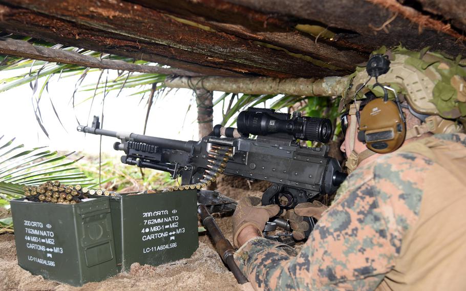 A Marine in combat gear and ear protection fires a machine gun from under cover in a foxhole.