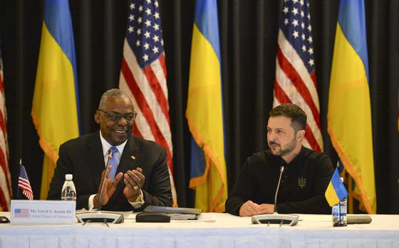 Lloyd Austin and Volodymyr Zelenskyy sit at a table in front of their countries’ flags.