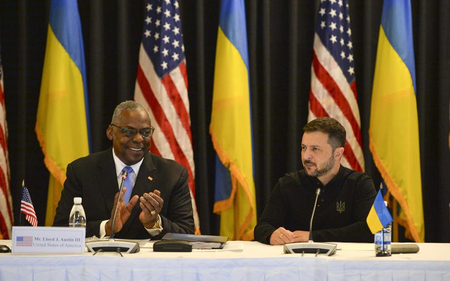 Lloyd Austin and Volodymyr Zelenskyy sit at a table in front of their countries’ flags.