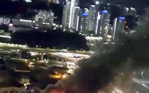 Smoke from a warehouse fire rises into the night sky with a city skyline in the background.