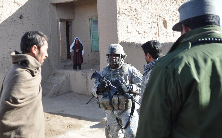 Staff Sgt. Darvin Cosby, 29, of Chicago, center,  stands with an Afghan National Police colleague, right, and a suspect arrested by the ANP, left, as a female relative looks on and police search the suspect’s home for bomb-making materials.