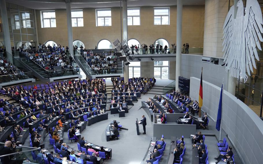 People fill seats in a semicircular great hall, while one person stands before all at a podium.