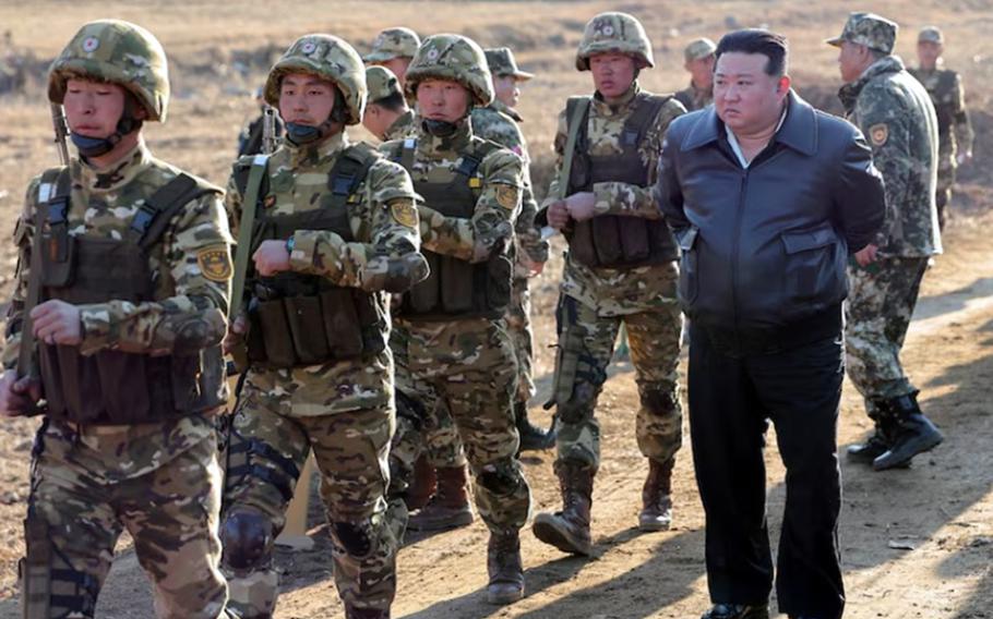 North Korean leader Kim Jong Un watches soldiers walking in a field wearing uniforms and vests.