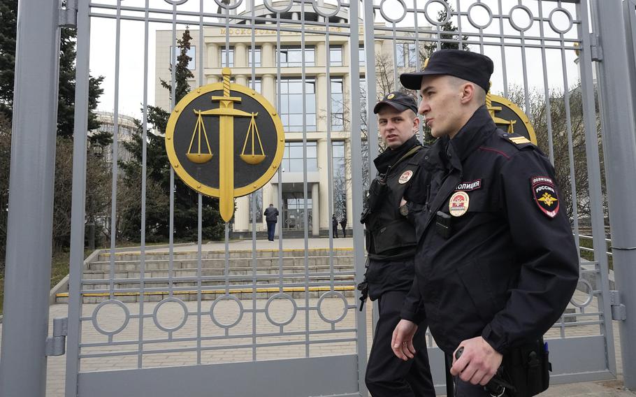 Russian police officers in uniform stand in front of a gate decorated with the scales of justice.