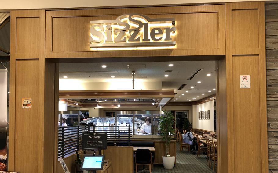 A restaurant interior can be seen from just outside a broad entrance flanked by wooden panels and a lit sign above.