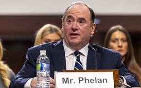 John Phelan, wearing a dark suit, sits behind a placard reading “Mr. Phelan” while he speaks into a microphone in a Senate hearing room.