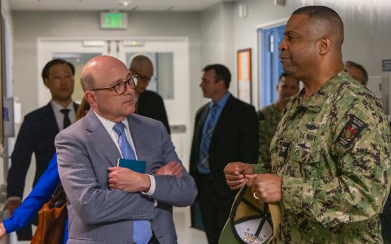 A bald man in glasses and a light grey suit speaks with a naval officer in camouflage uniform in the corridor of a hospital.