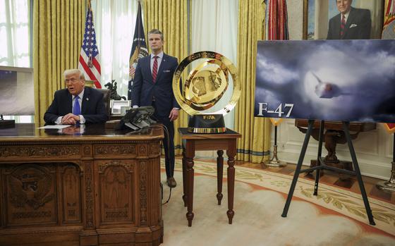 Donald Trump speaks while seated at a desk in the Oval Office and Pete Hegseth stands behind him with a photo of a airplane on an easel to their left.