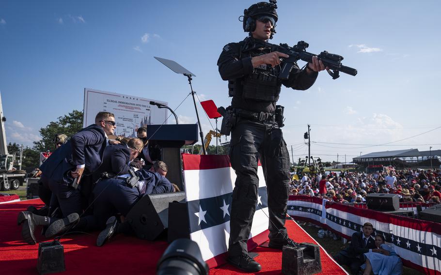 A Secret Service agent stands at the front of the stage, gun at the ready. Behind him,  agents surround and cover Donald Trump.