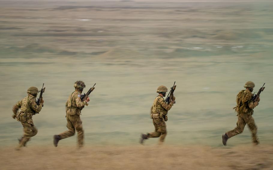 Four soldiers in combat gear and carrying machine guns run across a beach with water in the background.