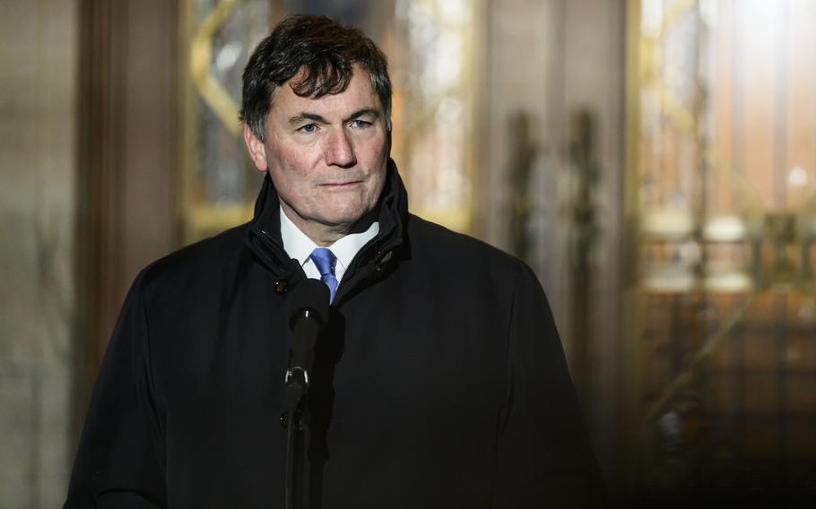 A man in a dark suit and coat speaks into a microphone during a press conference in front of a goverment building.