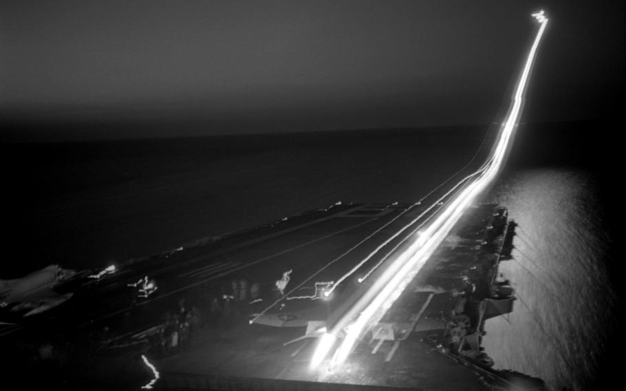 A black-and-white photo of a white trail stretching across a runway at sea and going into the sky.