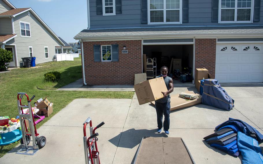 A man carries a cardboard box up a ramp into a moving truck on the edge of a driveway with an open garage door in the background.