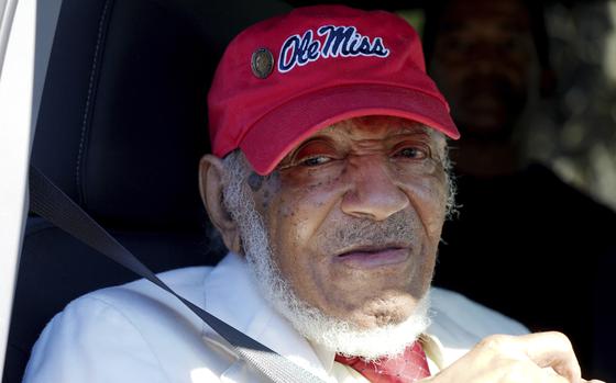 An elderly man in a red Ole Miss ball cap smiles slightly from his seat inside a car.