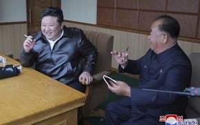 Kim Jong Un seated at a desk and smoking a cigarette while next to another seated official, apparently to inspect drone tests.