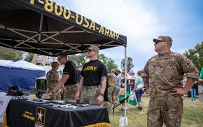 Soldiers stand near a table with informational pamphlets under a canopy at a recruitment booth.