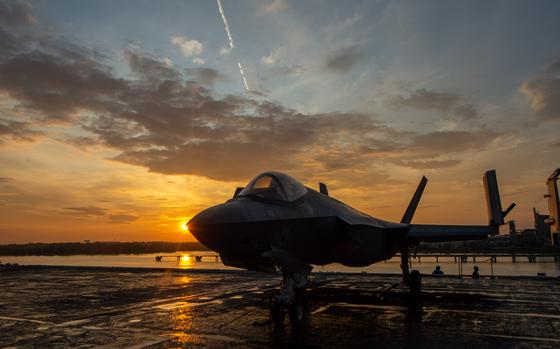 A fighter jet is parked on a flight deck with the sun low in the sky behind it.