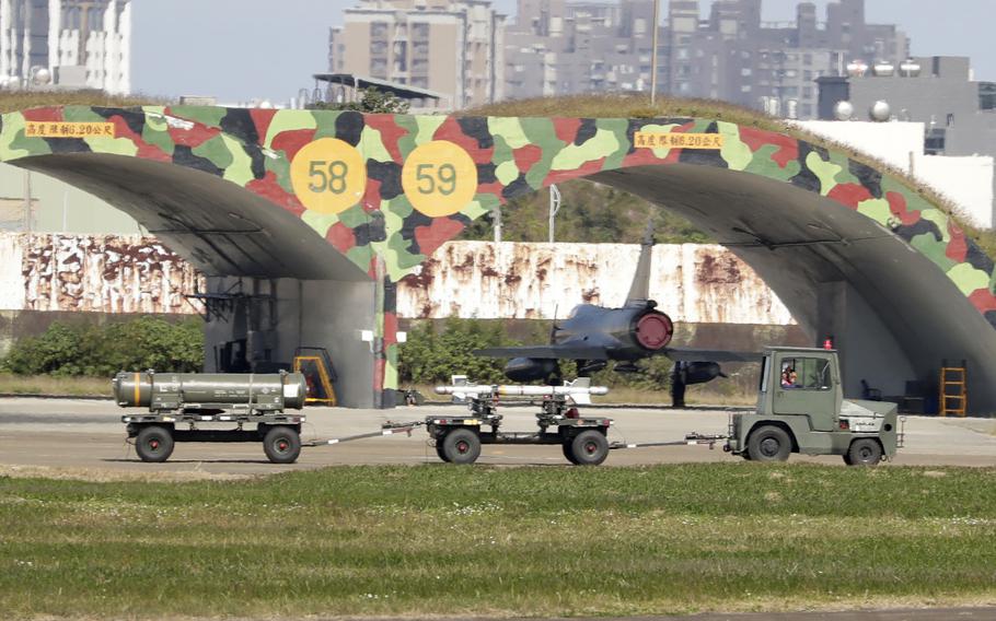 A military vehicle hauling rocket parts drives past a military airplane parked under a bridge.