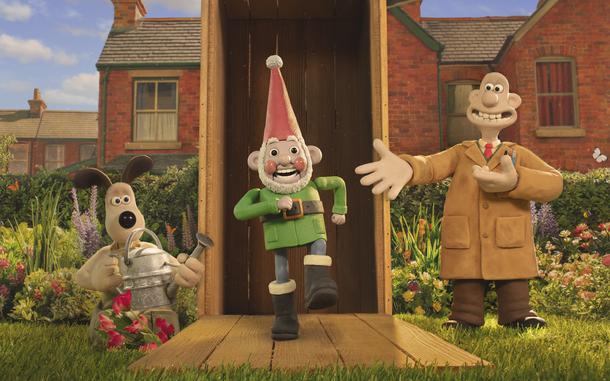 A stop-motion animation dog carrying a water can, a stop-motion animation gnome and a stop-motion animation human in front of a house in a garden.