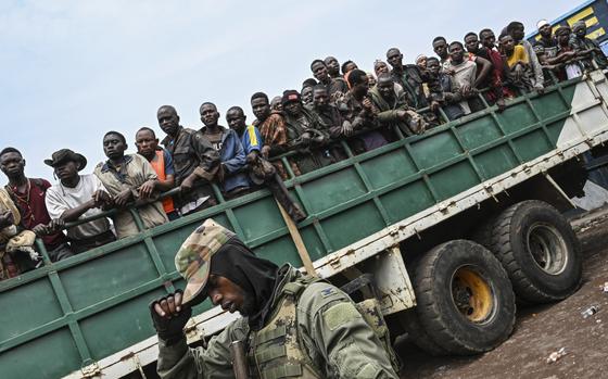 A group of men stand in the back of a flat-bed truck while an armed soldier in combat gear stands next to the truck.