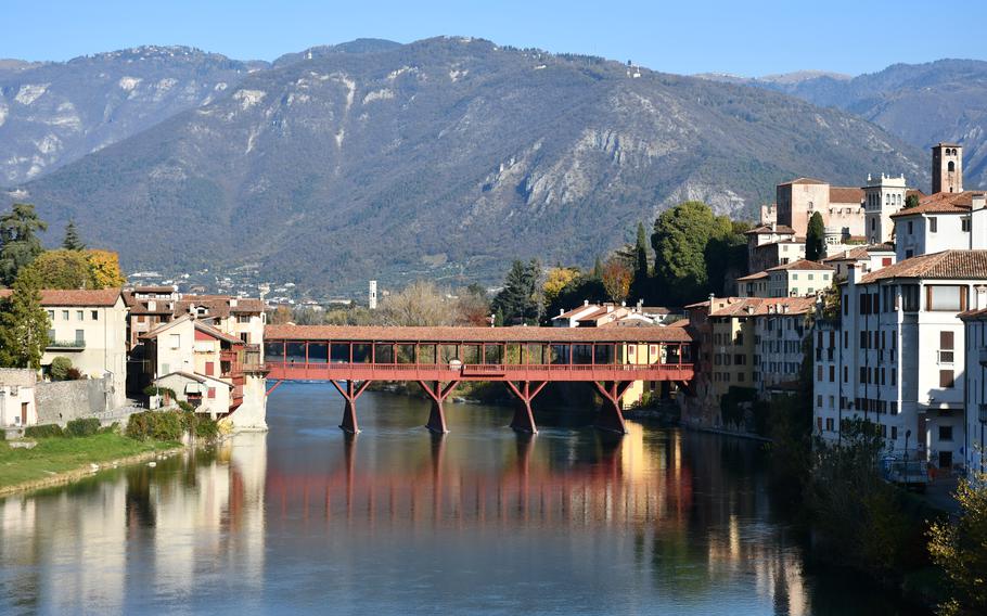 The Ponte Vecchio covered bridge, seen from afar, stretches across the Brenta River in Bassano del Grappa, Italy, uniting buildings on either side, with mountains in the background on a sunny day.