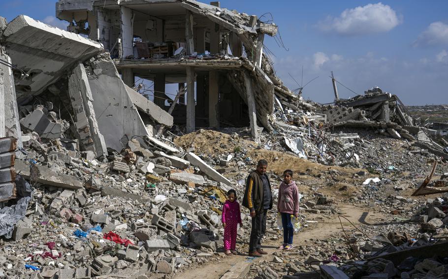 A man and two girls stand on a dirt path amid the rubble of destroyed homes.