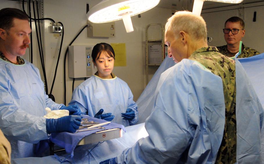 Military service members dressed in surgical scrubs and gloves stand over an operating table.