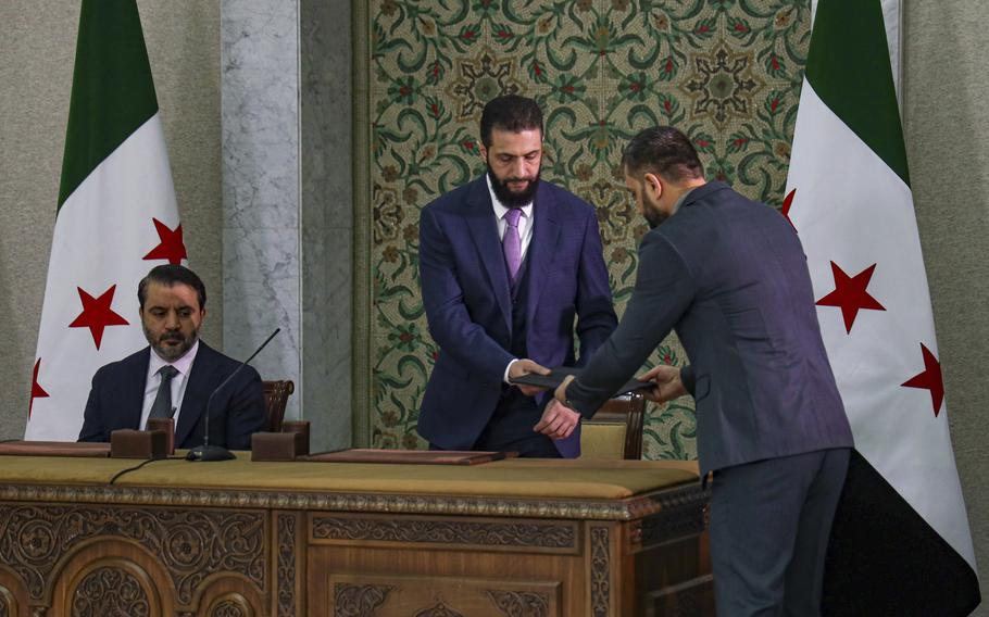 Syria’s interim president standing at an ornate desk, being handed a folder by another man.