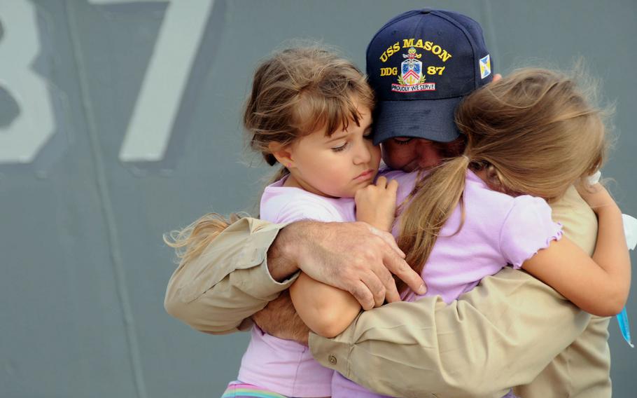 A sailor hugs his two young daughters while standing in front of a Navy destroyer.