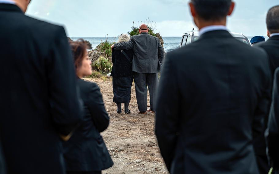 A man and a woman stand close together during a memorial for those killed in an Osprey crash. Other mourners are in the foreground.