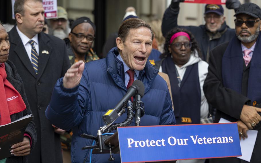 A man in a blue coat stands at a podium with a placard reading “Protect Our Veterans” and speaks into a microphone, with people standing behind him, some holding signs.