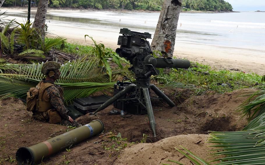 A Marine in combat gear sits in a trench among palm trees next to munitions on a tripod.