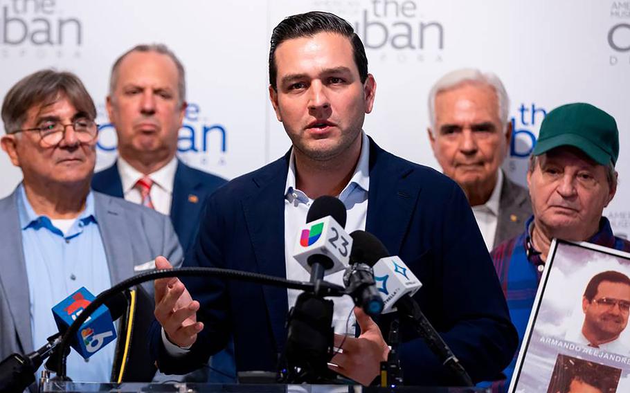 A man in a dark blazer stands at a podium in front of onlookers and speaks into a bank of microphones.