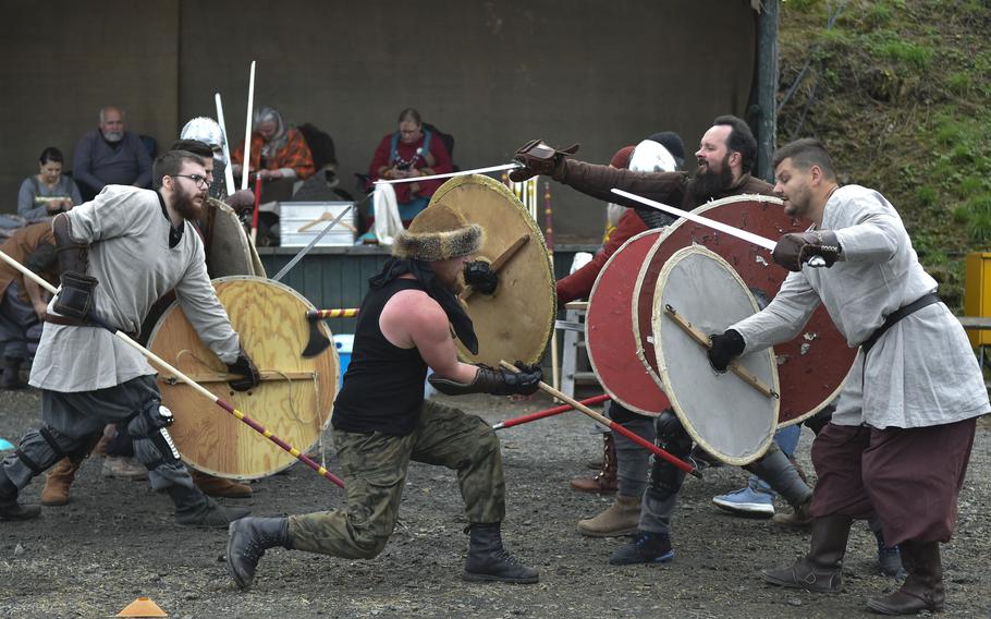 Reenactors wearing period garb strike each other with wooden swords and hold huge round wooden shields.