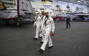 Three U.S. Navy sailors walk past aircraft inside a hangar on an aircraft carrier.