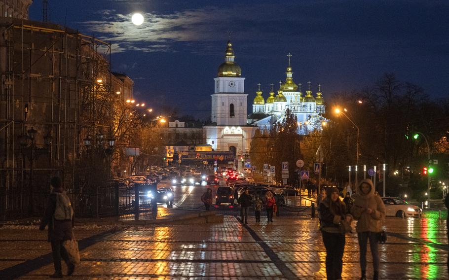 People walk on wet cobblestones at night in front of a golden roofed monastery in the background in Ukraine.