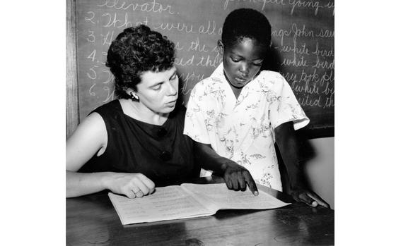 A black-and-white image of a teacher seated at a desk in front of a chalkboard, with a boy standing next to her pointing at a sheet of paper on the desk.