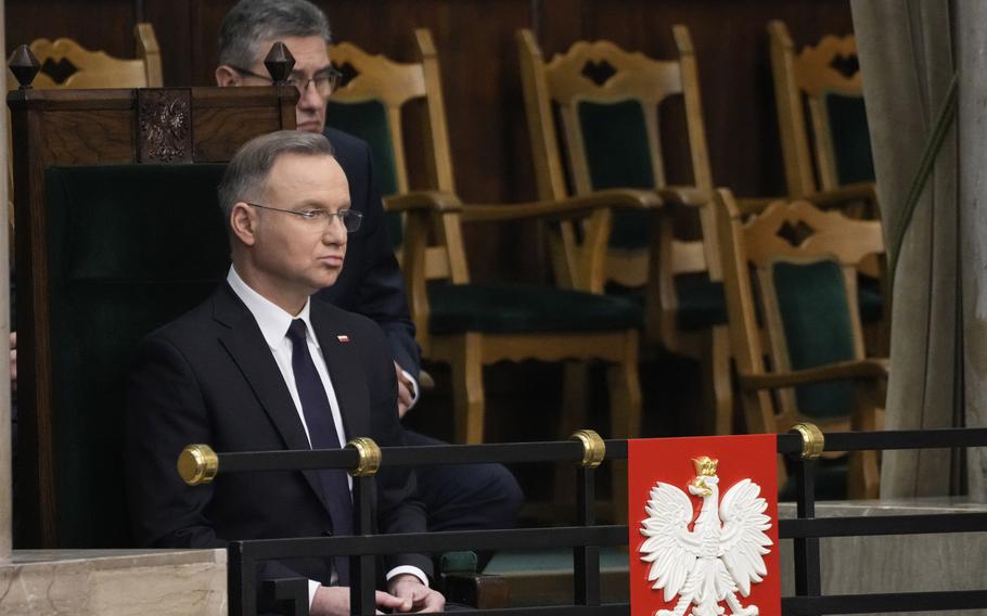 A man in a suit sitting in a seat with the Polish coat of arms on a railing in front of him.