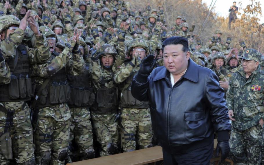 Kim Jong Un, clad in a black leather jacket and black pants, walks in front of a crowd of troops who are saluting.