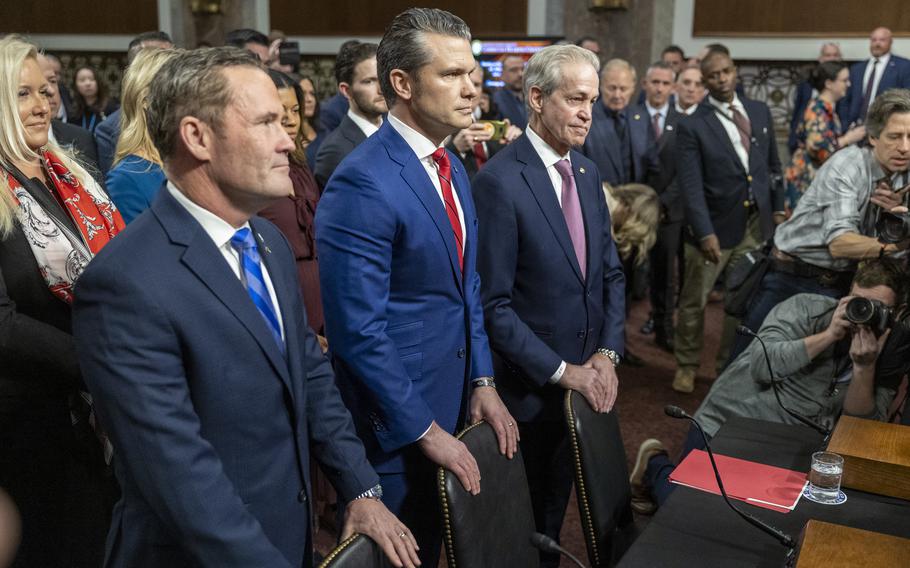 Three men in suits stand with their hands on the backs of their assigned seats.