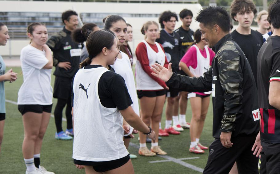 A Japanese soccer coach extends his hand to American high school players during a practice on a field.