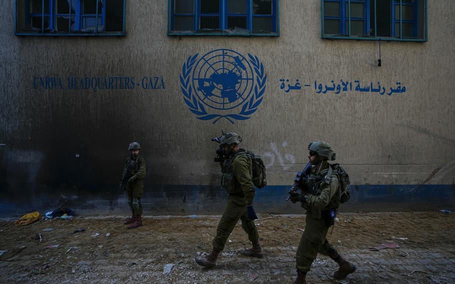 Israeli soldiers in uniform and holding guns walk in front of a damaged building with a symbol of the United Nations painted on the side.