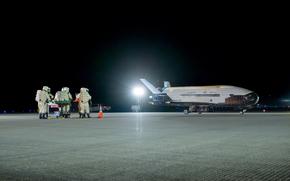 Personnel in white safety suits and helmets stand on the runway before a spacecraft.