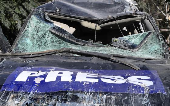 Close-up view of a destroyed car with “PRESS” painted on the hood with a shattered windshield and crumpled roof.