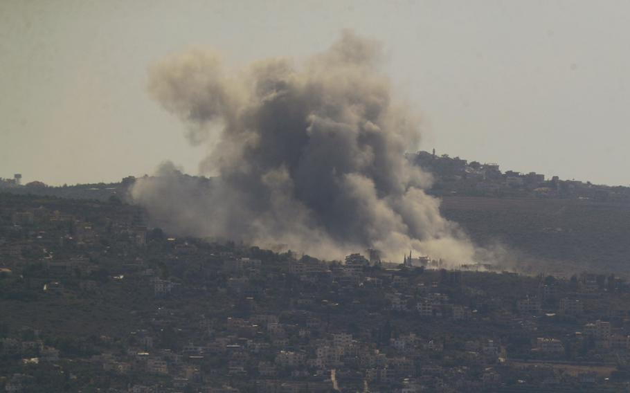 Smoke rises into the sky above the buildings of a village on a hill.