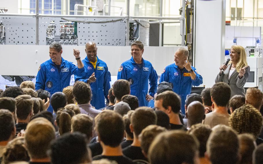 Four NASA astronauts in blue next to the SpaceX CEO in a suit, all of whom are in front of a crowd.