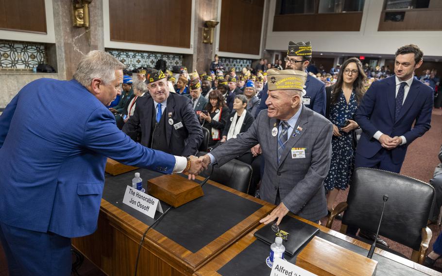 Bost leans over a table to shakes hands with a man wearing a veterans hat as others look on.